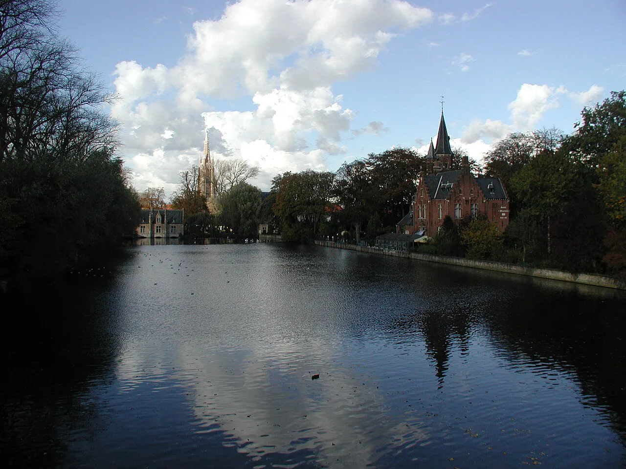 The Minnewater lake in Bruges with a church spire rising beyond the trees.